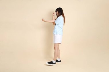 Açık mavi polo tişört giyen genç bir Asyalı kadın, ve beyaz şort, düz bir bej arka planda armache. Üniversite öğrencisi, Gen Z, Summer