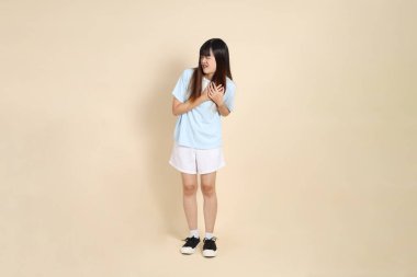 Açık mavi polo tişört ve beyaz şort giyen genç Asyalı bir kadın bej arka planda göğüs ağrısı çekiyor. Üniversite öğrencisi, Gen Z, Summer