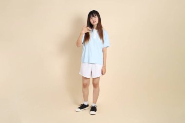 Açık mavi polo tişörtlü, beyaz şortlu genç bir Asyalı kadın bej arka planda sıcak hissediyor. Üniversite öğrencisi, Gen Z, Summer