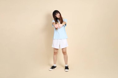 Açık mavi polo tişörtlü, beyaz şortlu genç bir Asyalı kadın bej arka planda egzersiz yapıyor. Üniversite öğrencisi, Gen Z, Summer