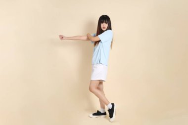 Açık mavi polo tişörtlü, beyaz şortlu genç bir Asyalı kadın bej arka planda egzersiz yapıyor. Üniversite öğrencisi, Gen Z, Summer