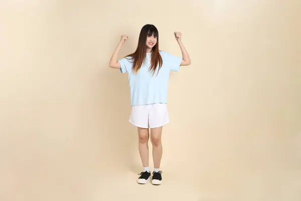 Açık mavi polo tişört giyen genç bir Asyalı kadın, ve beyaz şort, sade bej arka planda güçlü. Üniversite öğrencisi, Gen Z, Summer
