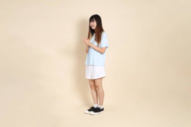 Açık mavi polo tişörtlü, beyaz şortlu, bej arka planda el çırpan genç Asyalı bir kadın. Üniversite öğrencisi, Gen Z, Summer