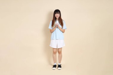 Açık mavi polo tişörtlü, beyaz şortlu, bej arka planda el çırpan genç Asyalı bir kadın. Üniversite öğrencisi, Gen Z, Summer