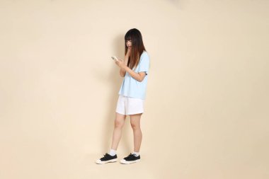 Açık mavi polo tişörtlü, beyaz şortlu, bej arka planda cep telefonu tutan genç bir Asyalı kadın. Üniversite öğrencisi, Gen Z, Summer