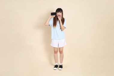 Açık mavi polo tişörtlü, beyaz şortlu, bej arka planda cep telefonu tutan genç bir Asyalı kadın. Üniversite öğrencisi, Gen Z, Summer