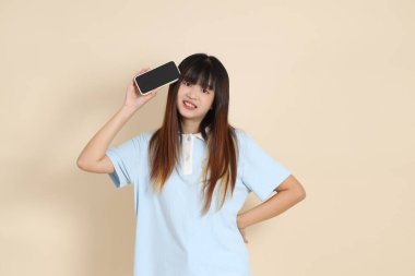 Açık mavi polo tişörtlü, beyaz şortlu, bej arka planda cep telefonu tutan genç bir Asyalı kadın. Üniversite öğrencisi, Gen Z, Summer