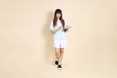 Açık mavi polo tişörtlü, beyaz şortlu, bej arka planda tablet tutan genç bir Asyalı kadın. Üniversite öğrencisi, Gen Z, Summer