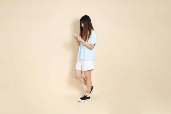 Açık mavi polo tişörtlü, beyaz şortlu, bej arka planda cep telefonu tutan genç bir Asyalı kadın. Üniversite öğrencisi, Gen Z, Summer