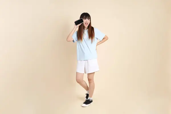 Açık mavi polo tişörtlü, beyaz şortlu, bej arka planda cep telefonu tutan genç bir Asyalı kadın. Üniversite öğrencisi, Gen Z, Summer