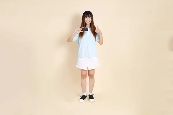 Açık mavi polo tişörtlü, beyaz şortlu, bej arka planda kredi kartı tutan genç bir Asyalı kadın. Üniversite öğrencisi, Gen Z, Summer