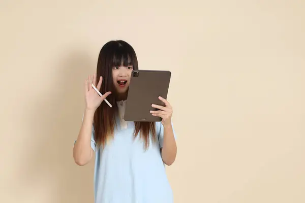 Açık mavi polo tişörtlü, beyaz şortlu, bej arka planda tablet tutan genç bir Asyalı kadın. Üniversite öğrencisi, Gen Z, Summer