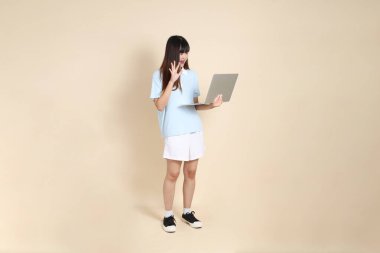 Açık mavi polo tişörtlü, beyaz şortlu, dizüstü bilgisayarı bej arka planda tutan genç bir Asyalı kadın. Üniversite öğrencisi, Gen Z, Summer
