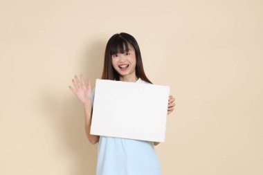 Açık mavi polo tişörtlü, beyaz şortlu, bej arka planda boş tuval tutan Asyalı genç bir kadın. Üniversite öğrencisi, Gen Z, Summer