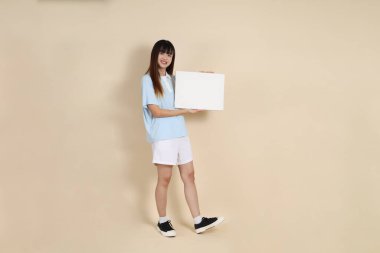 Açık mavi polo tişörtlü, beyaz şortlu, bej arka planda boş tuval tutan Asyalı genç bir kadın. Üniversite öğrencisi, Gen Z, Summer