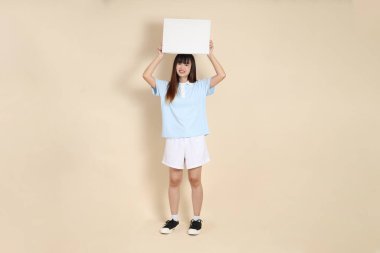 Açık mavi polo tişörtlü, beyaz şortlu, bej arka planda boş tuval tutan Asyalı genç bir kadın. Üniversite öğrencisi, Gen Z, Summer