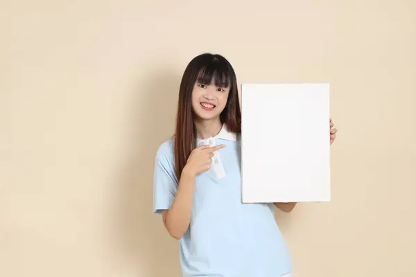 Açık mavi polo tişörtlü, beyaz şortlu, bej arka planda boş tuval tutan Asyalı genç bir kadın. Üniversite öğrencisi, Gen Z, Summer