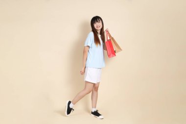 Açık mavi polo tişörtlü genç bir Asyalı kadın ve beyaz şortlu. Elinde bej arka planda bir alışveriş çantası var. Üniversite öğrencisi, Gen Z, Summer