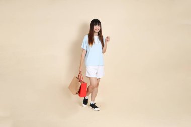 Açık mavi polo tişörtlü genç bir Asyalı kadın ve beyaz şortlu. Elinde bej arka planda bir alışveriş çantası var. Üniversite öğrencisi, Gen Z, Summer