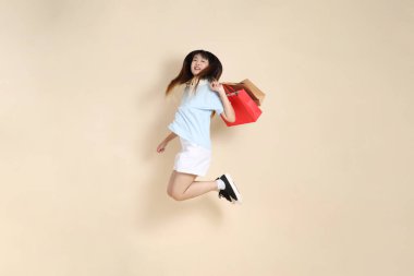 Açık mavi polo tişörtlü genç bir Asyalı kadın ve beyaz şortlu. Elinde bej arka planda bir alışveriş çantası var. Üniversite öğrencisi, Gen Z, Summer