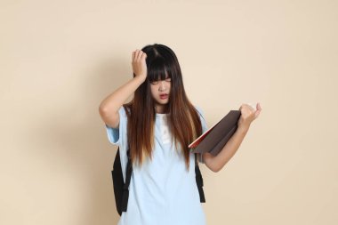 Asyalı bir kadın mavi polo tişört giyiyor, beyaz şort giyiyor, okul çantası taşıyor, elinde bej arka planda bir kitap tutuyor. Gen Z,