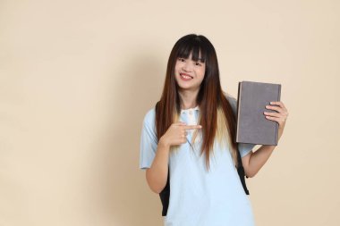 Asyalı bir kadın mavi polo tişört giyiyor, beyaz şort giyiyor, okul çantası taşıyor, elinde bej arka planda bir kitap tutuyor. Gen Z,