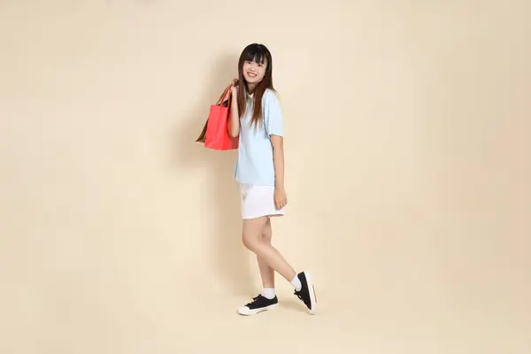 Açık mavi polo tişörtlü genç bir Asyalı kadın ve beyaz şortlu. Elinde bej arka planda bir alışveriş çantası var. Üniversite öğrencisi, Gen Z, Summer