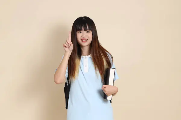 Asyalı bir kadın mavi polo tişört giyiyor, beyaz şort giyiyor, okul çantası taşıyor, elinde bej arka planda bir kitap tutuyor. Gen Z,