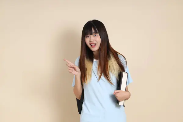 Asyalı bir kadın mavi polo tişört giyiyor, beyaz şort giyiyor, okul çantası taşıyor, elinde bej arka planda bir kitap tutuyor. Gen Z,