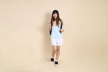 Asyalı bir kadın mavi polo tişört giyiyor, beyaz şort giyiyor, okul çantası taşıyor, elinde bej arka planda bir kitap tutuyor. Gen Z,