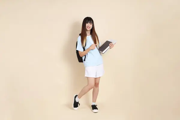 Asyalı bir kadın mavi polo tişört giyiyor, beyaz şort giyiyor, okul çantası taşıyor, elinde bej arka planda bir kitap tutuyor. Gen Z,
