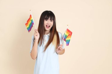 Açık mavi polo tişörtlü ve beyaz şortlu genç bir Asyalı kadın bej arka planda Gurur Bayrakları taşıyor. Üniversite öğrencisi, Gen Z, Summer