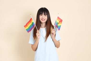 Açık mavi polo tişörtlü ve beyaz şortlu genç bir Asyalı kadın bej arka planda Gurur Bayrakları taşıyor. Üniversite öğrencisi, Gen Z, Summer