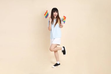 Açık mavi polo tişörtlü ve beyaz şortlu genç bir Asyalı kadın bej arka planda Gurur Bayrakları taşıyor. Üniversite öğrencisi, Gen Z, Summer