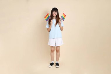 Açık mavi polo tişörtlü ve beyaz şortlu genç bir Asyalı kadın bej arka planda Gurur Bayrakları taşıyor. Üniversite öğrencisi, Gen Z, Summer