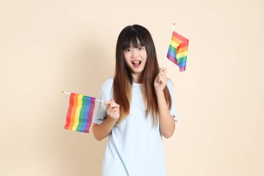 Açık mavi polo tişörtlü ve beyaz şortlu genç bir Asyalı kadın bej arka planda Gurur Bayrakları taşıyor. Üniversite öğrencisi, Gen Z, Summer