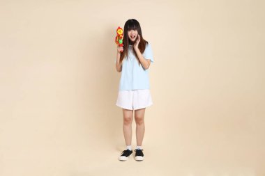 Açık mavi polo tişörtlü, beyaz şortlu, bej arka planda plastik su tabancası tutan genç bir Asyalı kadın. Üniversite öğrencisi, Gen Z, Summer