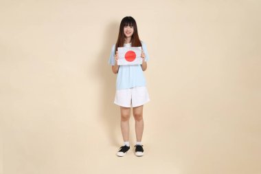 Açık mavi polo tişörtlü genç bir Asyalı kadın ve bej arka planda Japon ya da Japon bayrağı tutan beyaz şortlu. Üniversite öğrencisi, Gen Z, Summer
