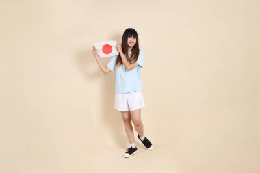 Açık mavi polo tişörtlü genç bir Asyalı kadın ve bej arka planda Japon ya da Japon bayrağı tutan beyaz şortlu. Üniversite öğrencisi, Gen Z, Summer