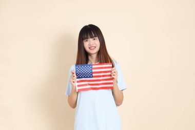 Açık mavi polo tişörtlü genç bir Asyalı kadın ve beyaz şortlu, ABD ya da Amerika Birleşik Devletleri ya da Amerikan bayrağını bej arka planda tutuyor. Üniversite öğrencisi, Gen Z, Summer