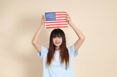 Açık mavi polo tişörtlü genç bir Asyalı kadın ve beyaz şortlu, ABD ya da Amerika Birleşik Devletleri ya da Amerikan bayrağını bej arka planda tutuyor. Üniversite öğrencisi, Gen Z, Summer