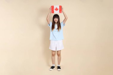 Açık mavi polo tişörtlü genç bir Asyalı kadın ve beyaz şortlu, bej arka planda Kanada ya da Kanada bayrağı taşıyor. Üniversite öğrencisi, Gen Z, Summer