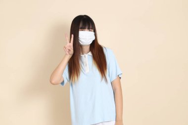 Sıradan kıyafetler giyen Asyalı bir kadın, sağlık maskesi takıyor. Covid-19 koruma işareti, V işareti, iki parmak, bej arka planda. Gen Z 