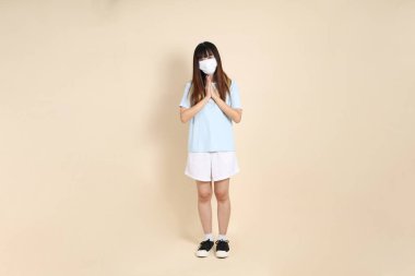 Sıradan kıyafetler giyen Asyalı bir kadın, tıbbi maske takıyor ve bej arka planda karşılama yapıyor. Z jenerasyonu, koruyucu solunum maskeleri..