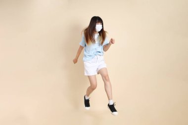 Sıradan kıyafetler giyen Asyalı bir kadın, tıbbi maske takıyor ve bej arka plana atlıyor. Z jenerasyonu, koruyucu solunum maskeleri..