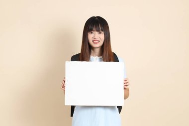 Asyalı bir kadın, mavi polo tişört giyiyor, beyaz şort giyiyor, okul çantası taşıyor, bej arka planda boş tuval tutuyor. Üniversite öğrencisi, Gen Z