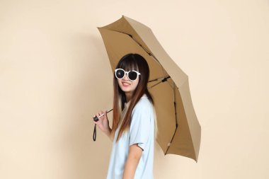 Mavi polo tişörtlü, beyaz şortlu, bej arka planda Şemsiye tutan Asyalı bir kadın. Üniversite öğrencisi, Gen Z