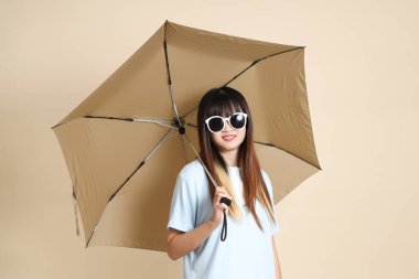 Mavi polo tişörtlü, beyaz şortlu, bej arka planda Şemsiye tutan Asyalı bir kadın. Üniversite öğrencisi, Gen Z