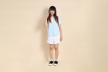 Açık mavi polo tişörtlü, beyaz şortlu, elinde düz bej bir kalem tutan genç Asyalı bir kadın. Üniversite öğrencisi, Gen Z, Summer