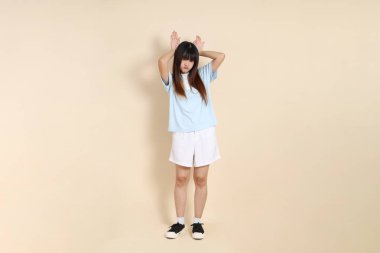 Orta boy Asyalı bir kadının açık mavi polo tişörtü ve beyaz şort giydiği fotoğraf. Sade bir bej arka plan karşısında aptalca ve komik. Üniversite öğrencisi, Gen Z